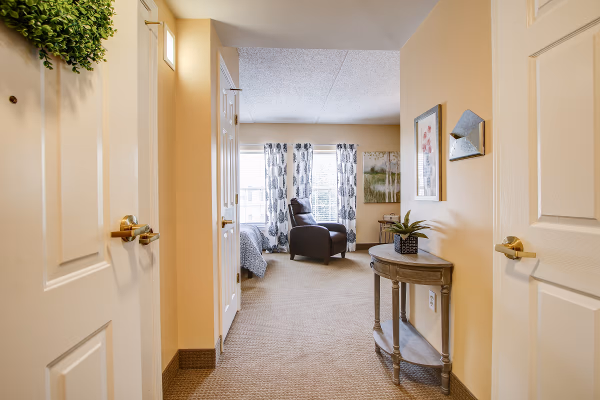View down a hallway into a bedroom with beige walls and carpet. The bedroom has a window with patterned curtains, a dark brown armchair, and a bed partially visible on the left. A small wooden table with a plant and wall art are on the right side of the hallway.