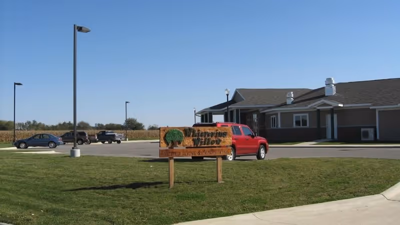 Exterior view of Whispering Willow facility with a wooden sign in the foreground that reads 'Whispering Willow Assisted Living & Memory Care'. The building is single-story with a gray roof and beige siding. Several cars are parked in the parking lot under a clear blue sky.