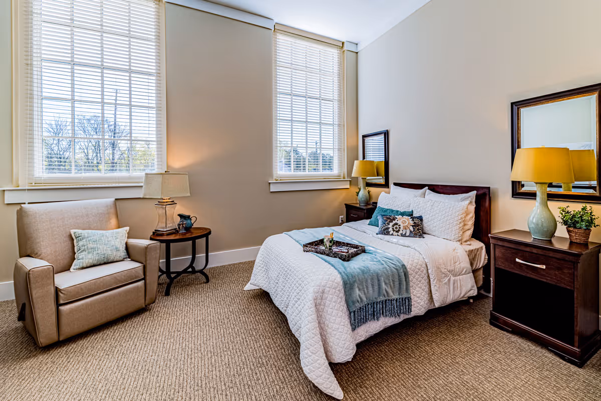 Bright furnished bedroom with a single bed, armchair, two nightstands with lamps, and large windows with blinds.