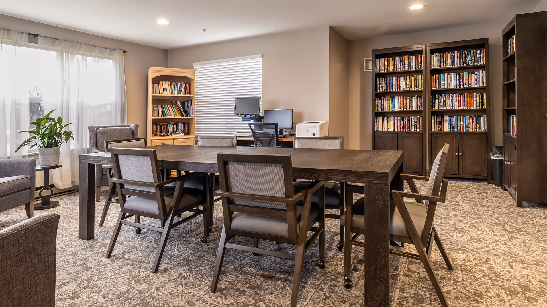 A well-lit room with a large rectangular wooden table surrounded by six cushioned chairs on wheels. The room features carpeted flooring with a patterned design, two tall bookshelves filled with books, a smaller bookshelf, a computer desk with a monitor and printer, and a potted plant near a window with sheer white curtains.