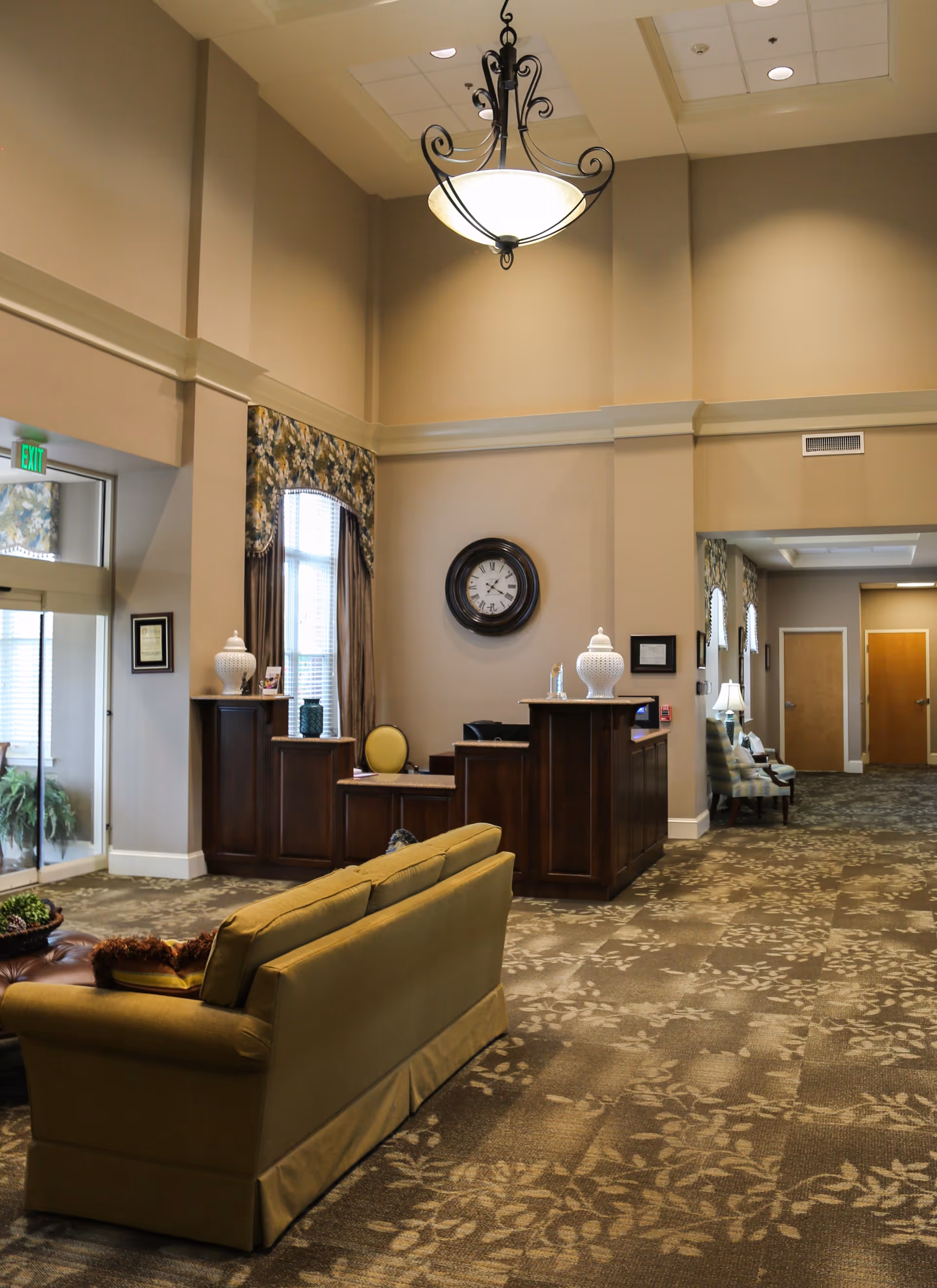 Spacious lobby with a sofa facing a wooden reception desk, wall clock, chandelier, and patterned carpet.