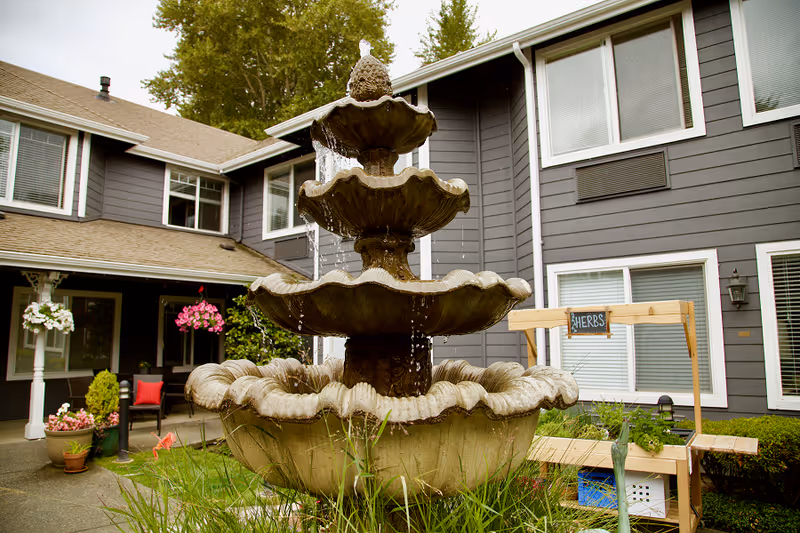 Outdoor courtyard area of a senior living facility with a large tiered water fountain in the center. Surrounding the fountain are green plants and flowers. In the background, there is a gray building with white-trimmed windows and a wooden stand labeled 'HERBS' holding potted plants. There are also hanging flower baskets and outdoor seating visible.