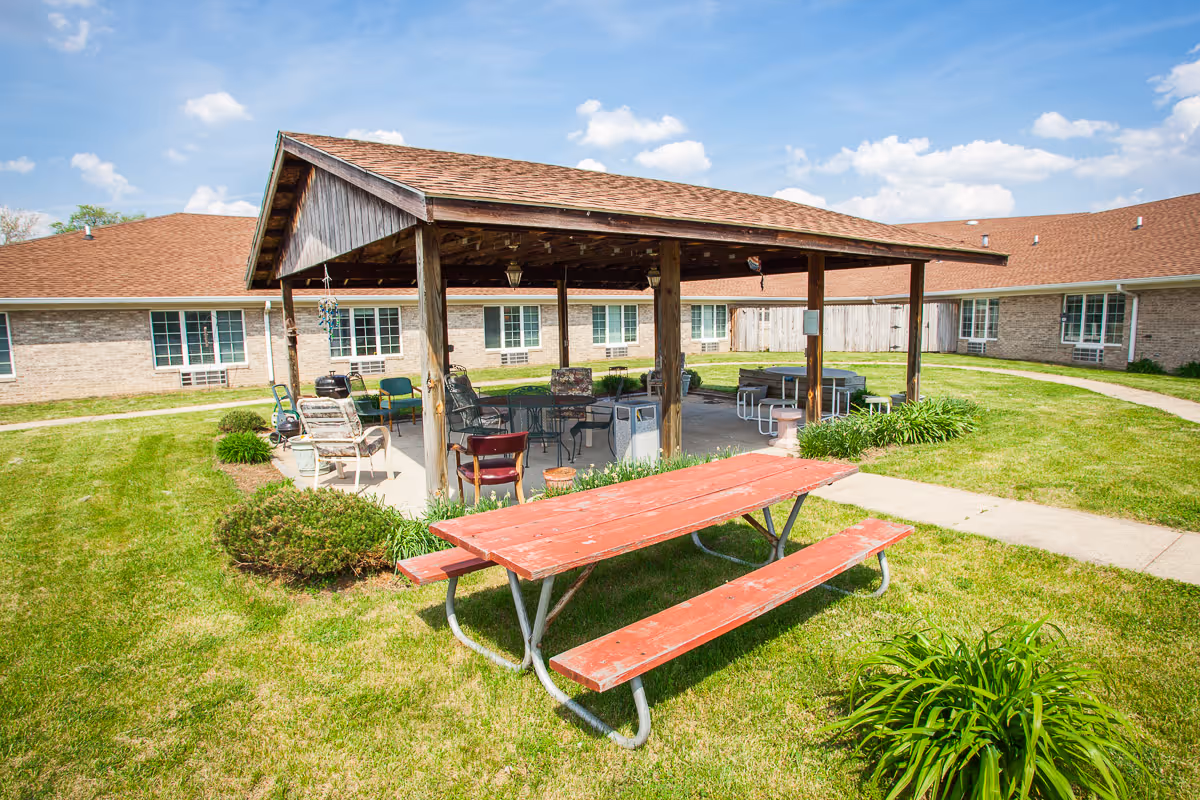 A grassy courtyard featuring a wooden pavilion with outdoor seating and a red picnic table in front of a single-story brick building.