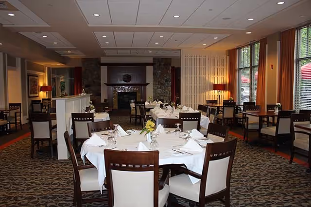 Dining room with round tables dressed in white linens and place settings, wooden chairs, carpeted floor, and a fireplace at the far end.
