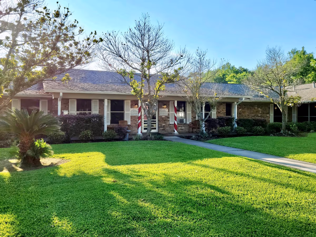 Single-story brick building with a well-maintained green lawn and several trees in front. The building has a covered porch with two chairs and two candy cane striped columns near the entrance. The sky is clear and blue.
