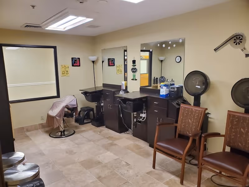 Interior view of a hair salon area in a senior living facility with two styling chairs in front of large mirrors, a hair washing station, two hair dryers mounted on the wall, and two brown cushioned chairs for waiting. The room has beige walls and tiled flooring.