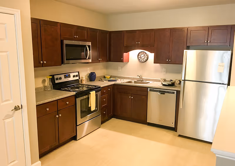 A modern kitchen with dark wood cabinets, stainless steel appliances including a refrigerator, oven, microwave, and dishwasher. The countertop has a few decorative items and a clock is mounted on the wall above the sink.