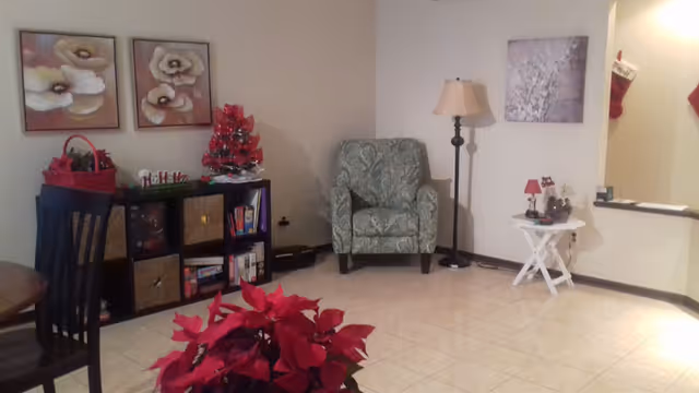 A cozy living room area with a patterned armchair, a floor lamp, a small white side table with decorative items, a shelving unit with baskets and books, and holiday decorations including a small red Christmas tree and poinsettia plants. The walls have floral artwork and there is a partial view of a dining table and chairs.