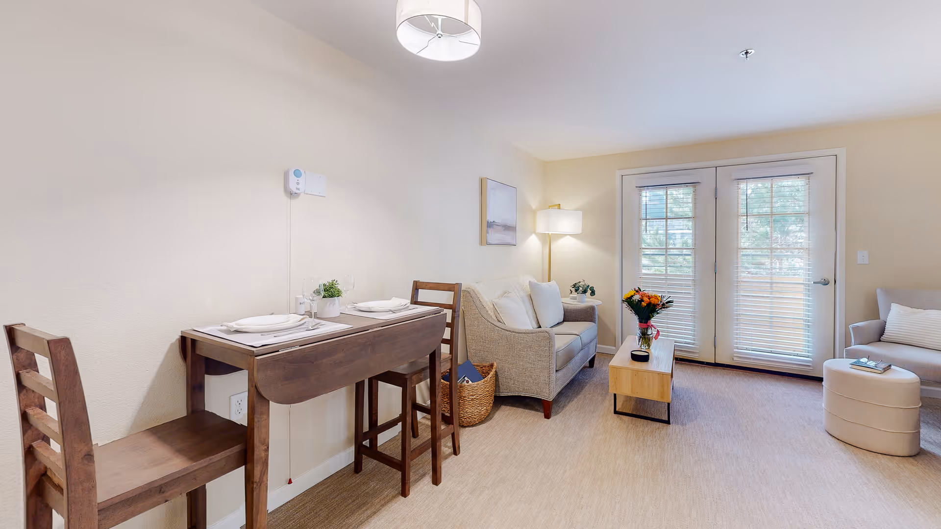Bright living room with a small drop-leaf dining table and chairs, a loveseat, coffee table with flowers, an armchair and French doors.