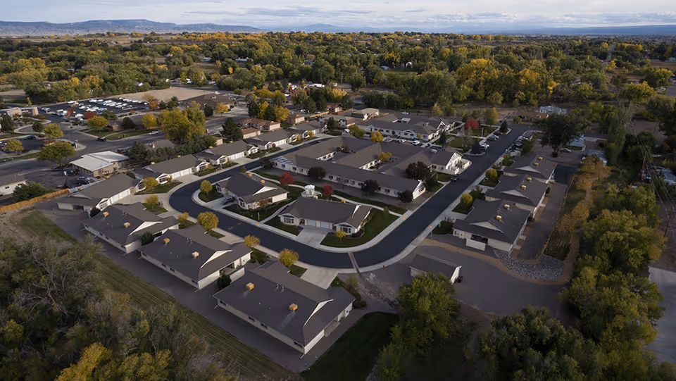 Aerial view of a senior living facility named Crossroads at Delta, showing multiple single-story buildings with gray roofs arranged around curved roads, surrounded by trees and greenery under a partly cloudy sky.