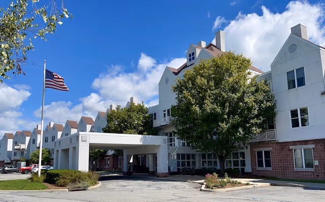 Exterior view of Glen Arden Inc, a multi-story senior living facility with white walls and brown roof accents. The building has multiple windows and a covered entrance area. There are trees and shrubs around the entrance, an American flag on a flagpole, and a partly cloudy blue sky above.