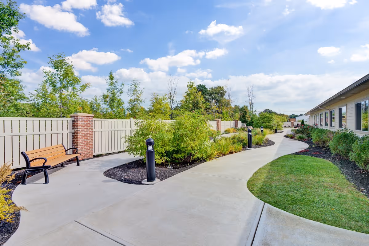 A curved concrete pathway in an outdoor garden area with green shrubs, trees, and a wooden bench along a white fence with brick pillars. The sky is blue with scattered clouds, and a building with windows is visible on the right side.