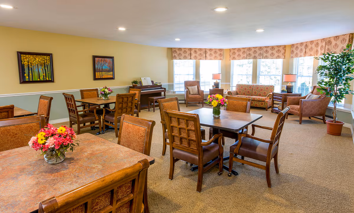 A bright and cozy common area in a senior living facility featuring several wooden tables with chairs, each table decorated with a vase of colorful flowers. The room has large windows with patterned valances allowing natural light to fill the space. There is a piano against one wall, two framed paintings, and a seating area with upholstered chairs and a patterned sofa near the windows. A potted plant and two table lamps add to the warm ambiance.
