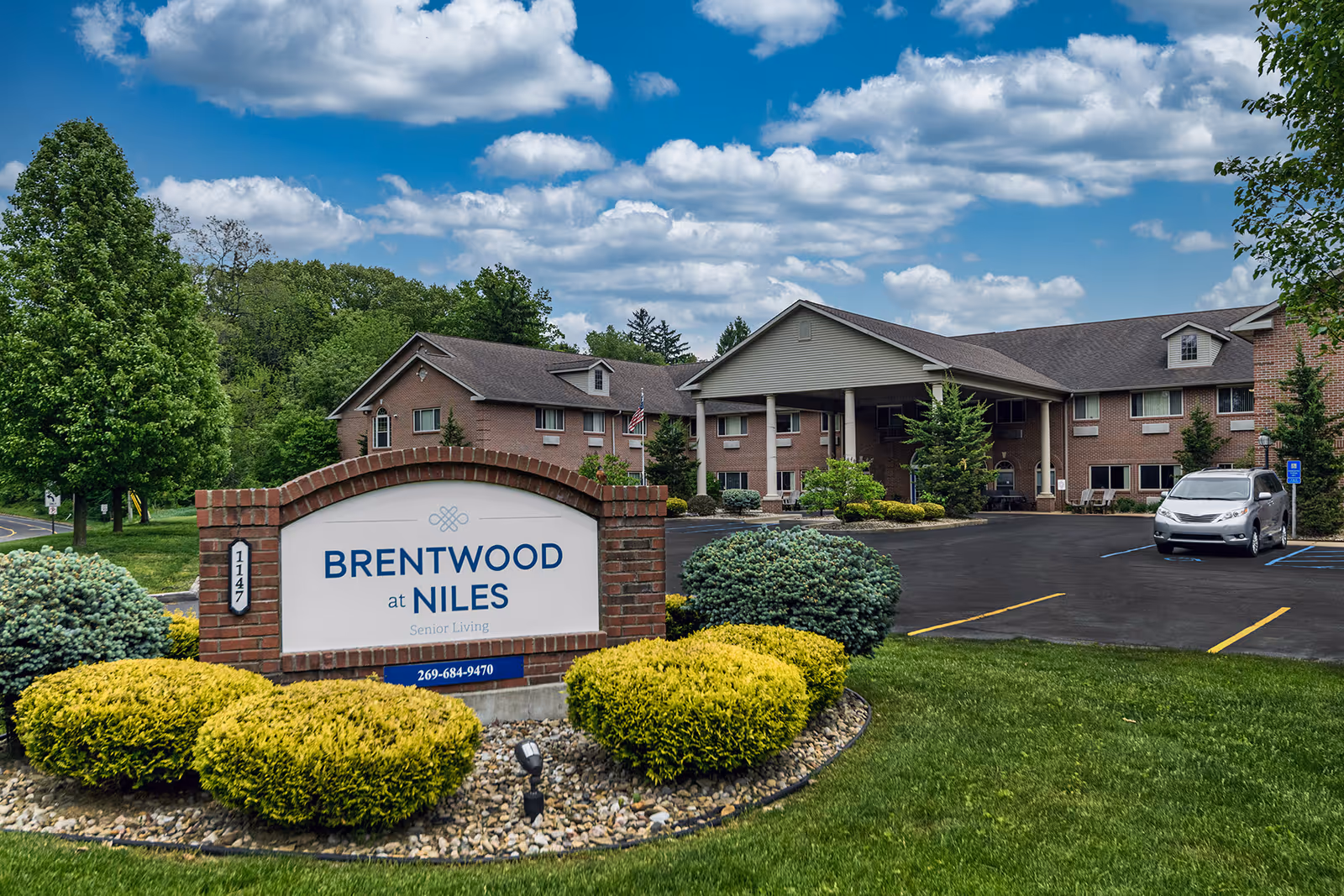 Exterior view of Brentwood at Niles Senior Living facility showing a brick building with a covered entrance, surrounded by green bushes and trees under a partly cloudy sky. A sign with the facility name and phone number is prominently displayed in the foreground.