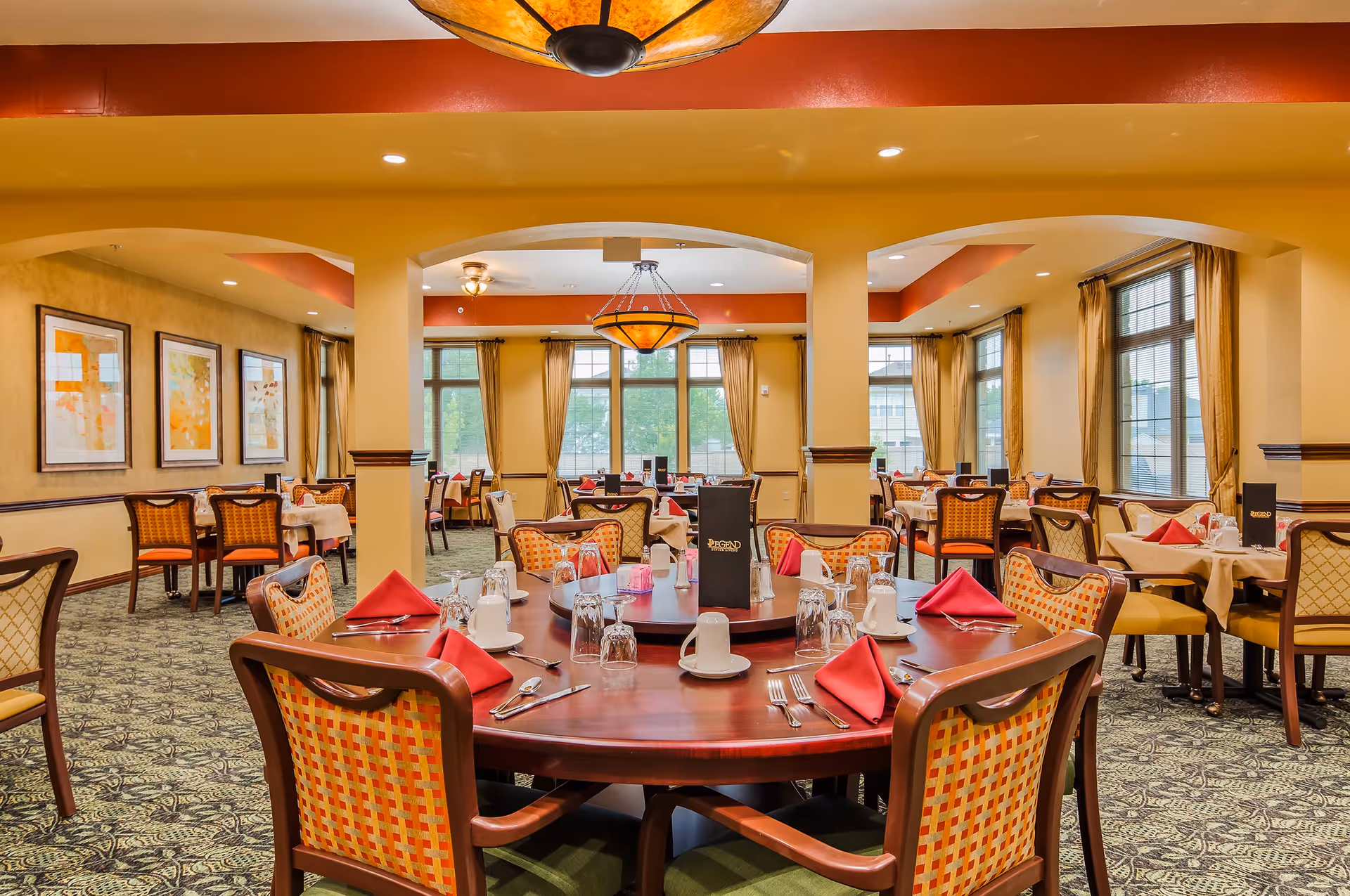 A spacious dining room with multiple tables set for a meal. The tables have red napkins, white cups, glasses, and silverware neatly arranged. The room features large windows with beige curtains, warm yellow walls, patterned carpet, and decorative ceiling lights. The central round table has a menu labeled 'Legend'.