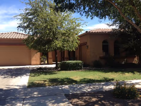 Front exterior of a single-story stucco building with a tiled roof, garage, driveway, trees, and a lawn.