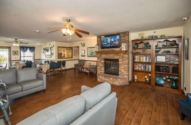 A spacious common area in a senior living facility featuring two gray sofas, a brick fireplace with a mounted flat-screen TV above it, a wooden bookshelf filled with books and decorative items, and a dining area with tables and chairs in the background. The room has wooden flooring, ceiling fans with lights, and framed artwork on the walls.