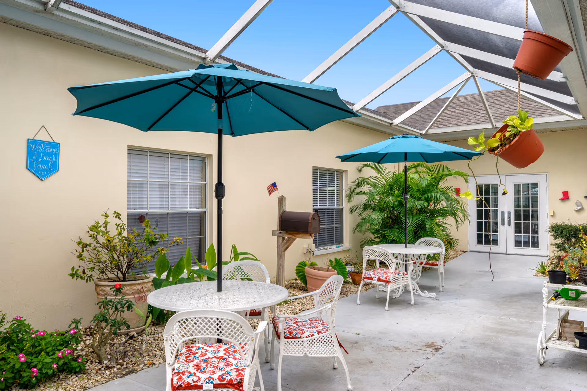 A bright and airy covered outdoor patio area with white metal tables and chairs featuring colorful cushions. Two teal umbrellas provide shade over the tables. Potted plants and greenery decorate the space, with a small mailbox and a blue sign on the wall that reads 'Welcome to the Back Porch.' The area is enclosed with glass panels overhead and has double glass doors at the far end.