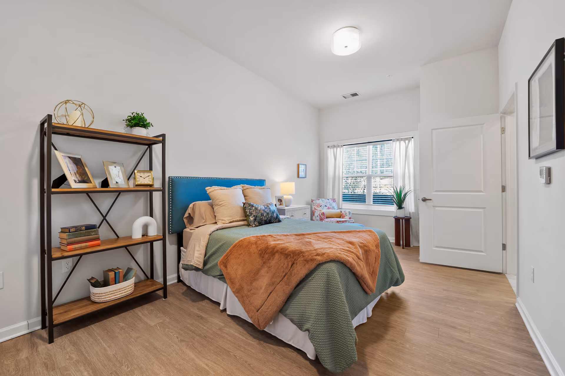 A bright and neatly arranged bedroom with a bed featuring a blue upholstered headboard, green bedding, and a brown throw blanket. To the left of the bed is a wooden bookshelf with decorative items and books. A window with white curtains lets in natural light, and a floral armchair sits near the window. The room has light wood flooring and white walls.