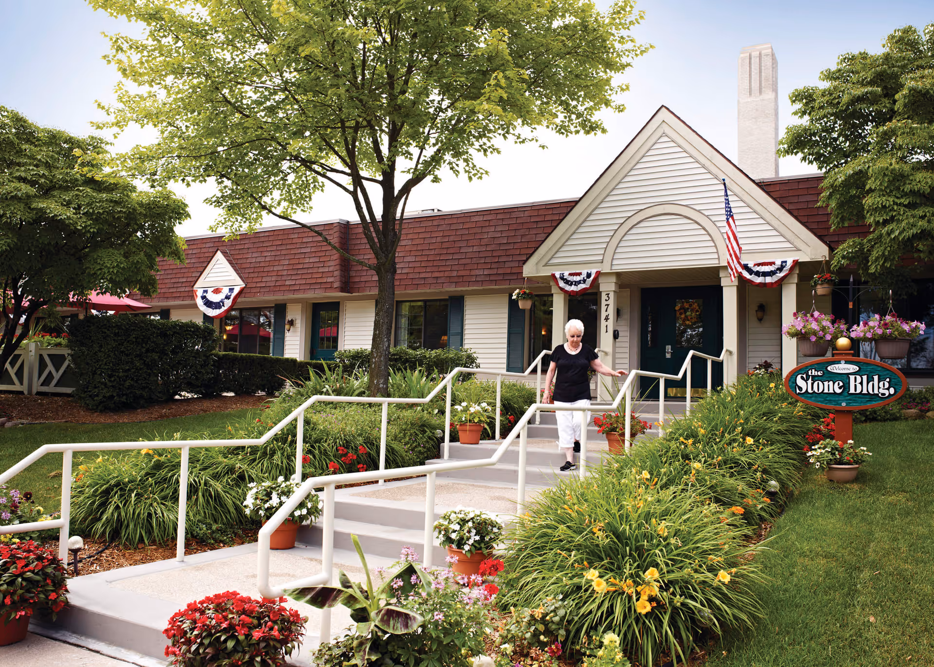 An elderly woman walking down a ramp with handrails outside a single-story building with a brown roof and white siding. The building has an American flag and patriotic bunting decorations near the entrance. There are well-maintained flower beds and green shrubs along the ramp and walkway. A sign near the entrance reads 'Welcome to the Stone Bldg.'