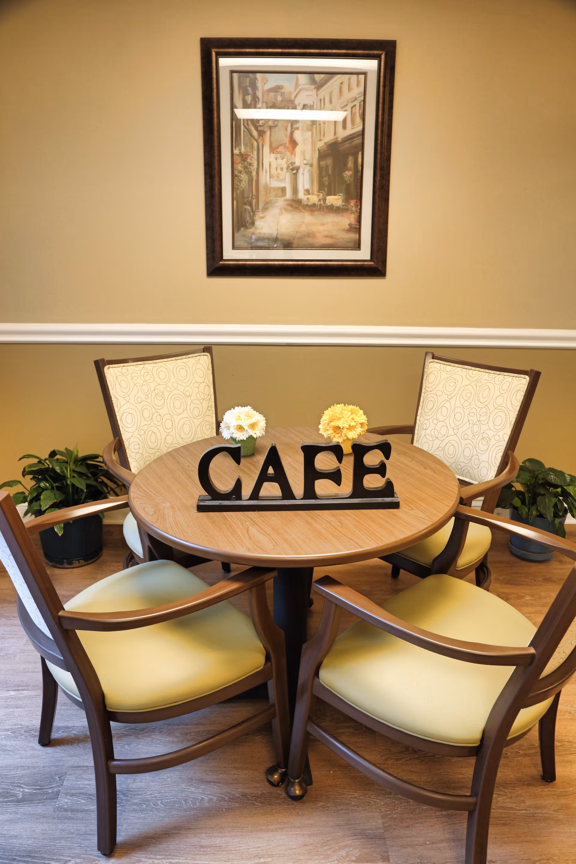 Round café table with a decorative 'CAFE' sign, four chairs, small flower arrangements and a framed painting on the wall.
