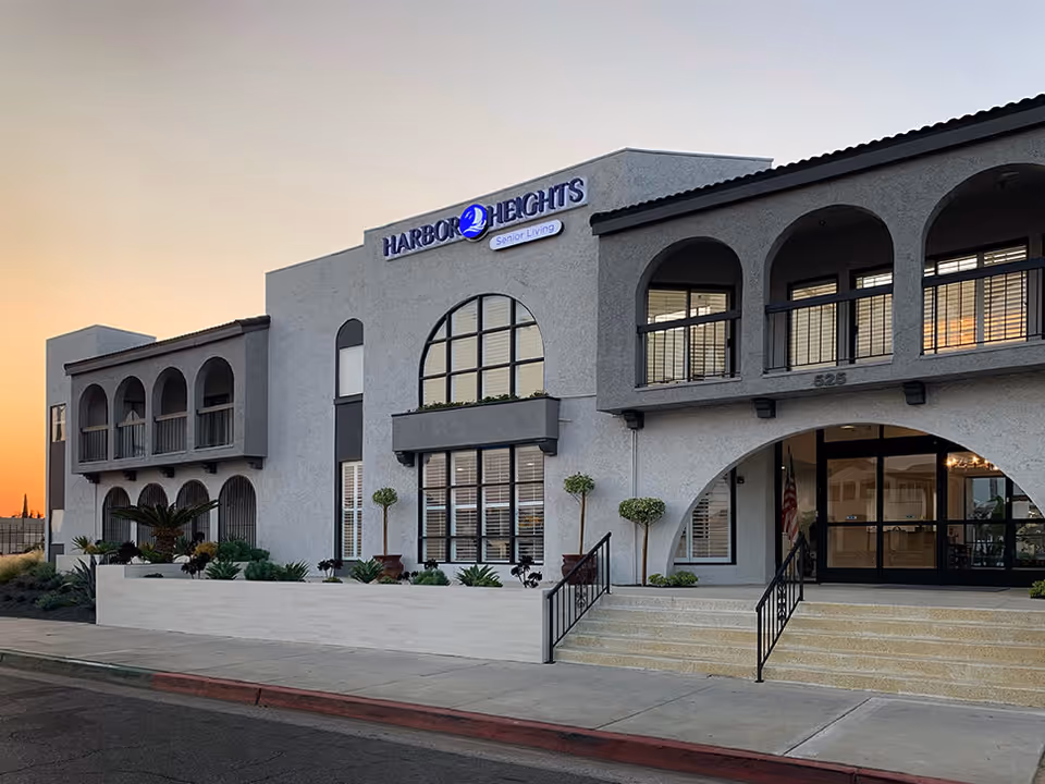 Exterior view of Harbor Heights senior living facility building at sunset, showing a two-story structure with arched windows and balconies, a staircase leading to the entrance, and landscaping with small trees and plants.