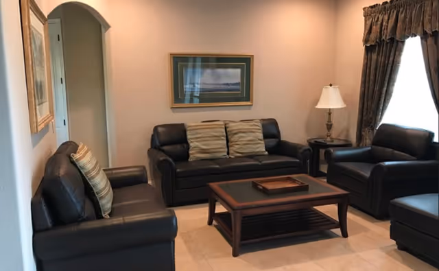 A cozy living room with three dark leather sofas arranged around a wooden coffee table. There are two striped cushions on the central sofa. A framed landscape painting hangs on the wall above the sofa. A side table with a lamp is next to one of the sofas, and a window with heavy curtains lets in natural light.