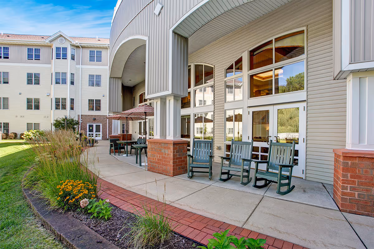 Outdoor patio at a senior living facility with rocking chairs, tables and umbrellas in front of large windows and landscaped grounds.