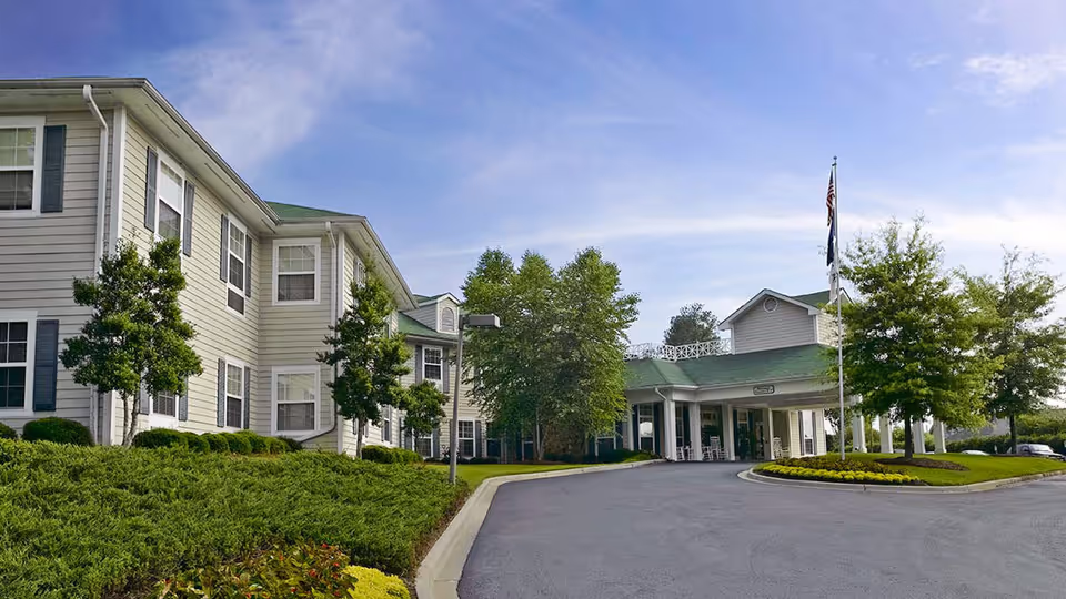 Exterior view of Forest Lake Manor, a two-story senior living facility with light-colored siding and green roofs. The entrance features a covered drop-off area with white columns, surrounded by well-maintained landscaping including trees, shrubs, and a circular flower bed with a flagpole displaying the American flag. The sky is clear with a few clouds.