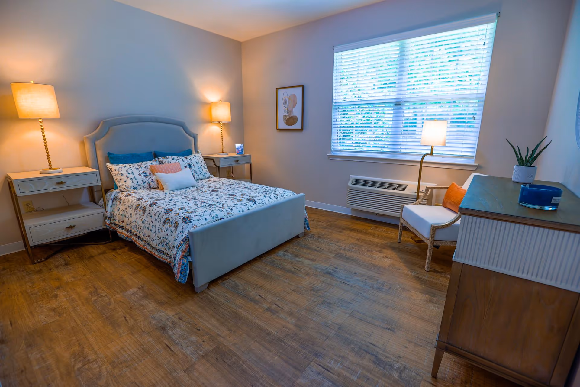 A cozy bedroom with a bed featuring a floral patterned comforter and multiple pillows. There are two bedside tables with lamps on either side of the bed. A window with blinds lets in natural light, and a chair with an orange cushion sits next to a floor lamp. A dresser with a plant and decorative items is also visible.