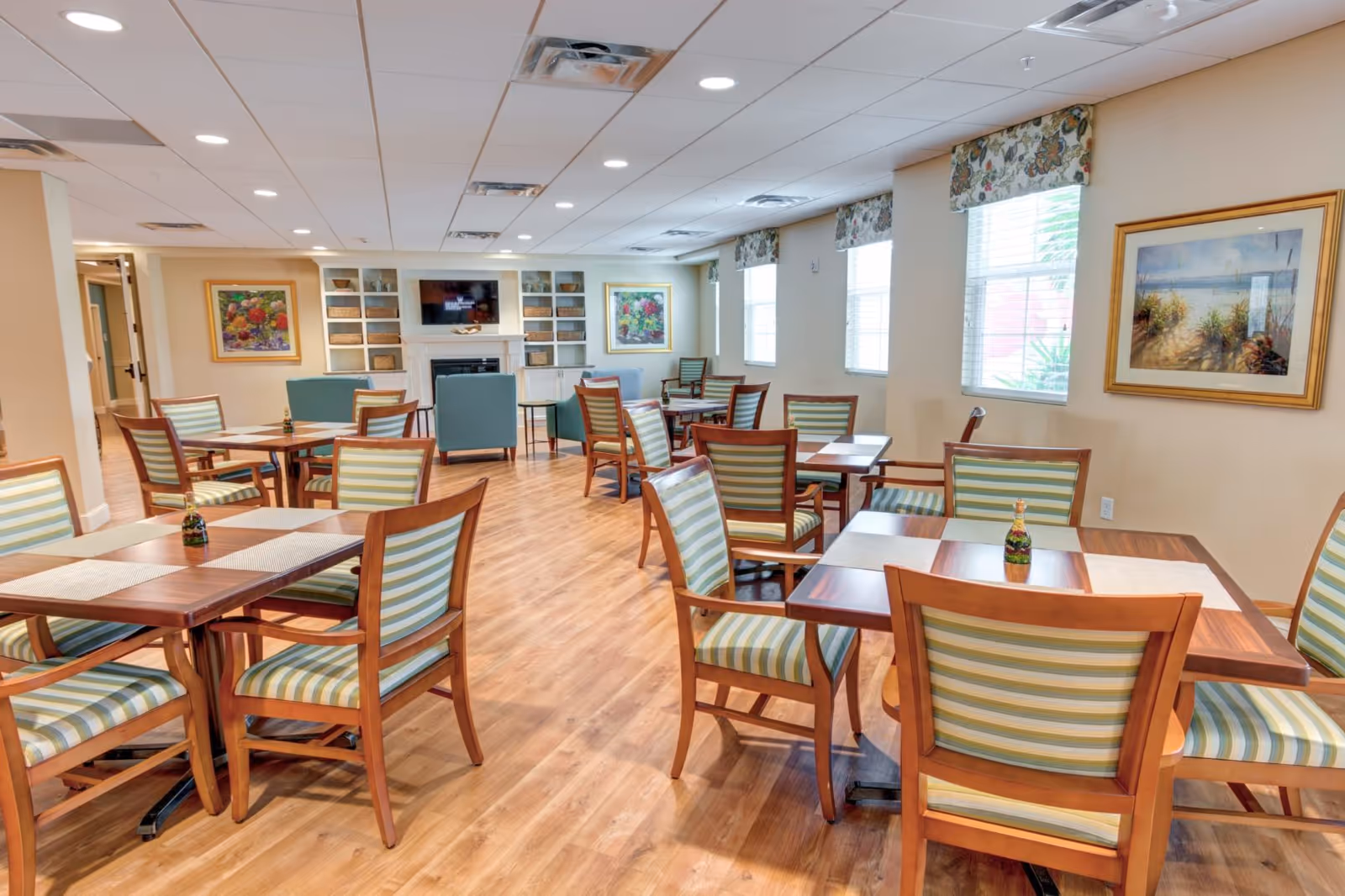 A bright dining room with multiple wooden tables and chairs featuring green and beige striped cushions. The room has wood flooring, large windows with floral valances, framed artwork on the walls, and a built-in shelving unit with a TV and fireplace at the far end.