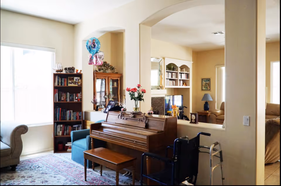A bright living room area with a wooden piano and matching bench in the center. A vase with pink flowers sits on top of the piano. To the left, there is a blue armchair and a bookshelf filled with books. A wheelchair and a walker are positioned near the piano. The room has beige walls, large windows with blinds, and an archway leading to another seating area with a lamp and more furniture.