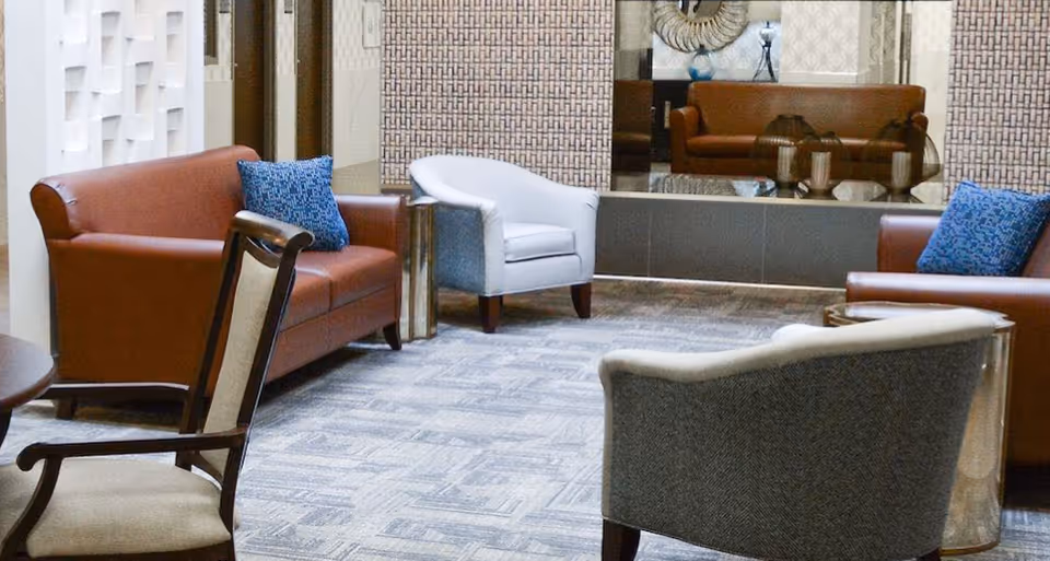 A furnished lounge with leather sofas, upholstered armchairs, blue pillows, and a decorative mirror on the far wall.