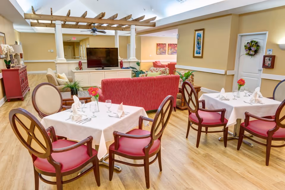 Open dining and lounge area in a senior living facility with set tables, red chairs, sofas, and a TV.