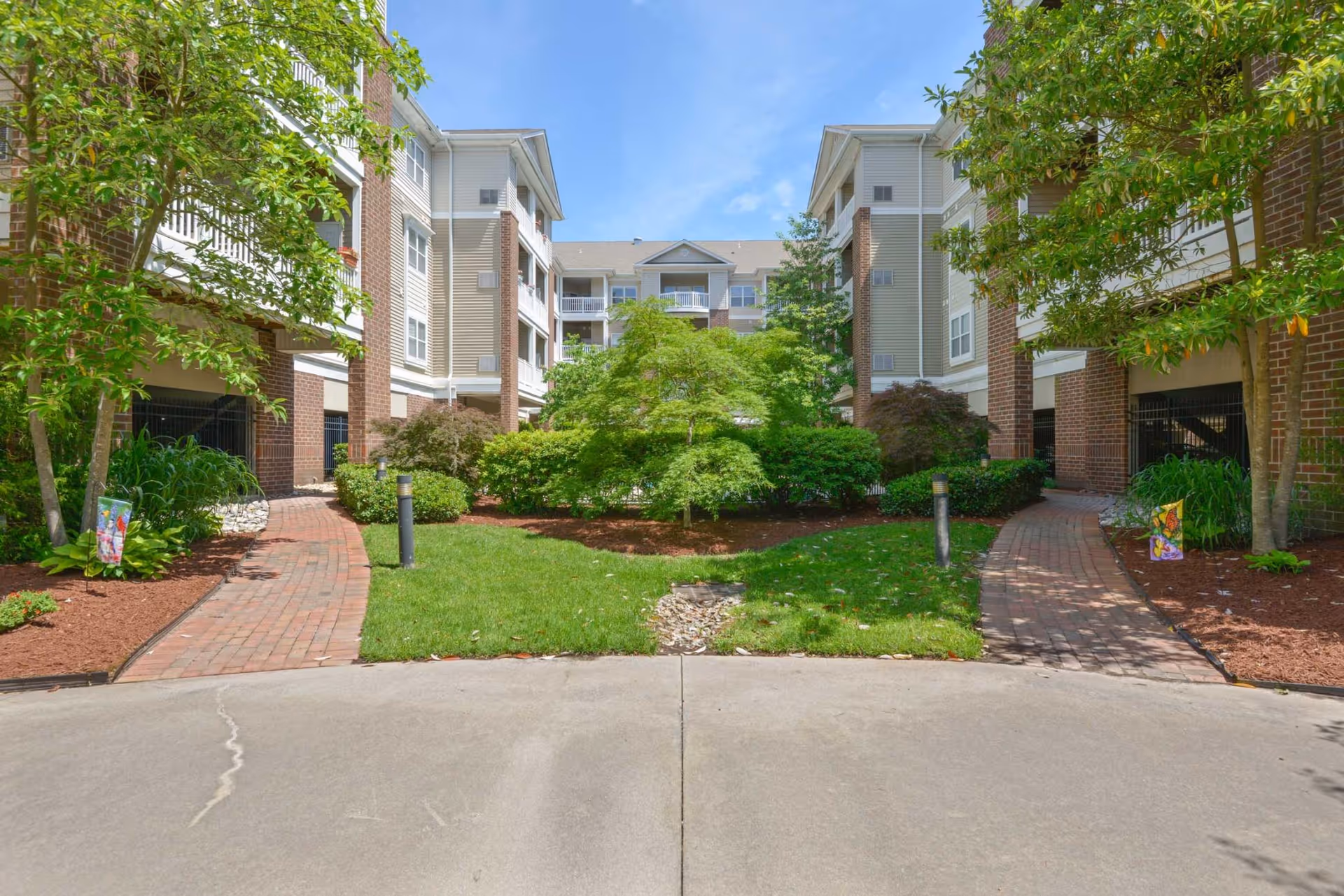 Landscaped courtyard with brick paths and greenery between two wings of a multi-story residential building.