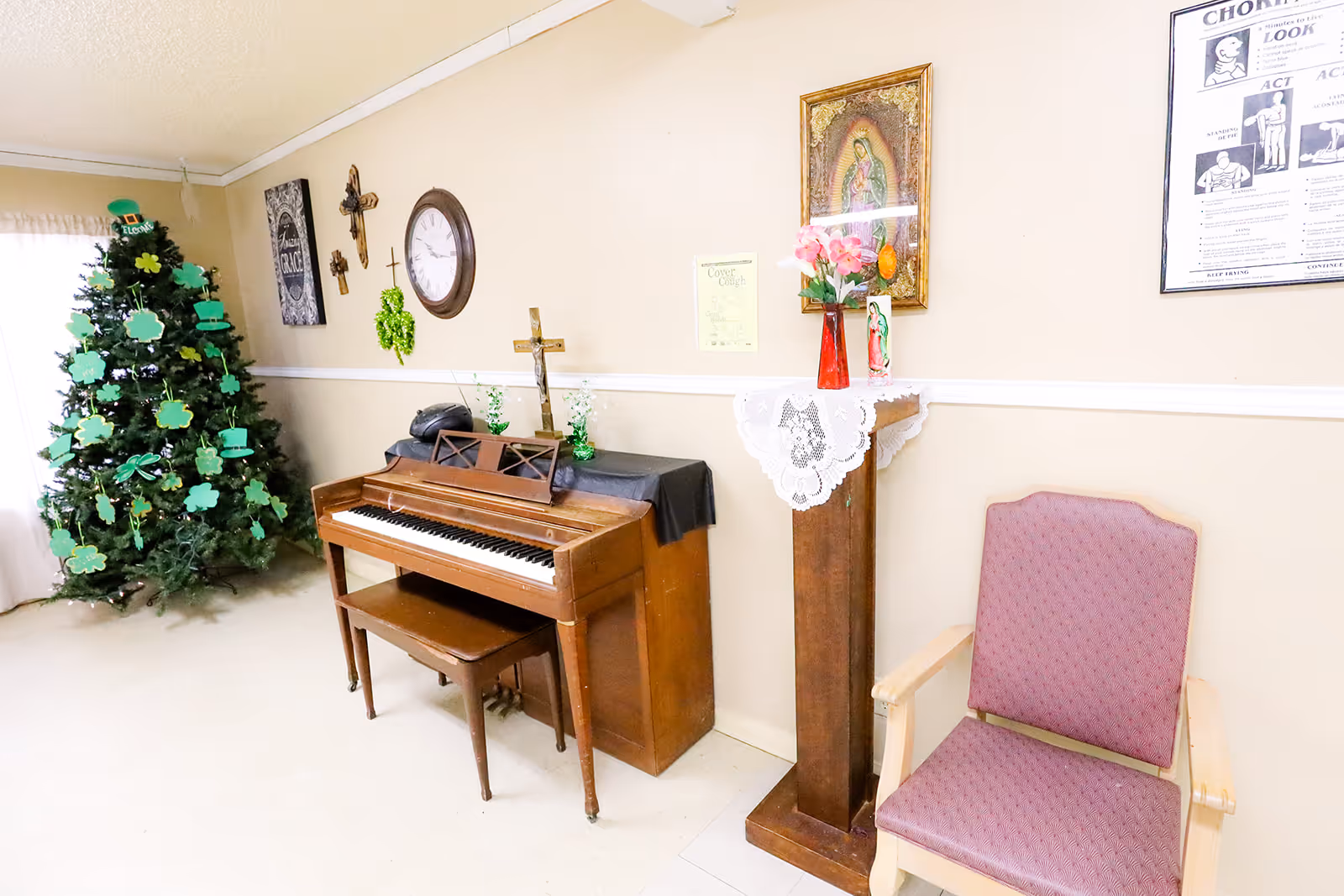 Interior common room with an upright piano, decorated Christmas tree, wall clock and crucifix, a pedestal with flowers, and a cushioned armchair.