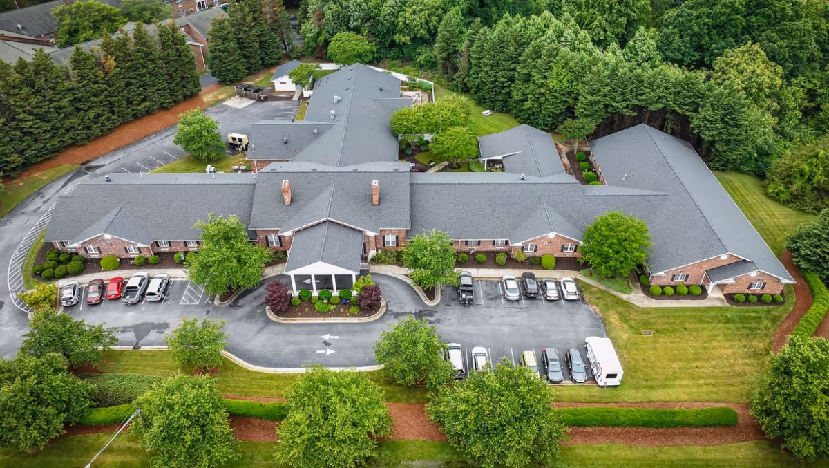 Aerial view of Kerner Ridge Assisted Living facility showing a large single-story brick building with a gray roof surrounded by green trees and landscaped grounds. Several cars are parked in the parking lot in front of the building.