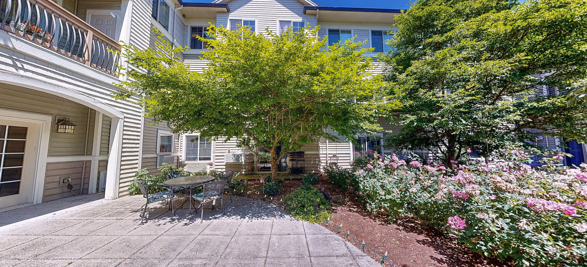 Outdoor patio area at Brookdale Vancouver Orchards featuring a round metal table with four chairs on a paved surface, surrounded by lush greenery including trees and flowering bushes. The building exterior with windows and balconies is visible in the background under a clear blue sky.