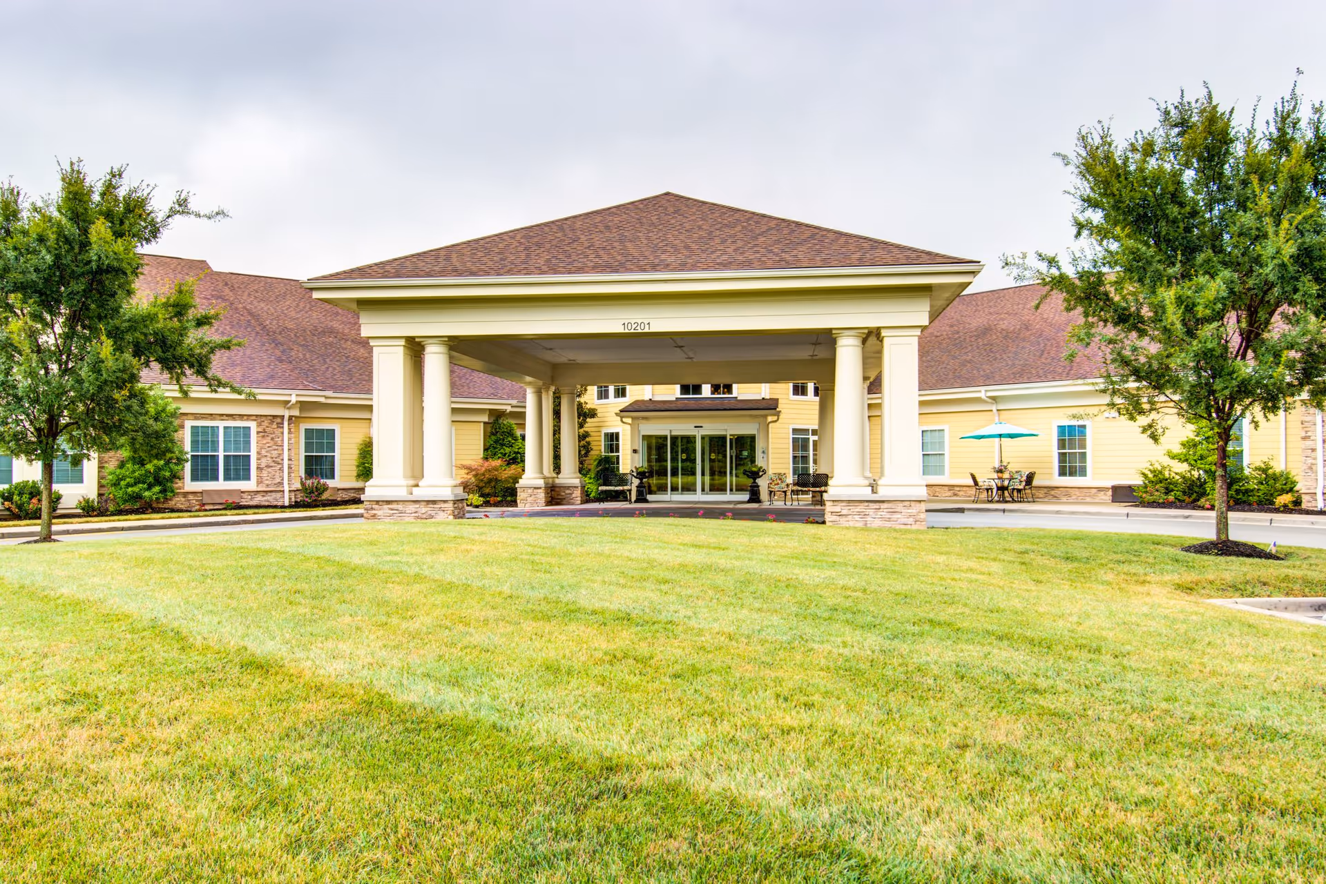 Sunny exterior entrance with a covered porte-cochere, lawn, and outdoor seating.