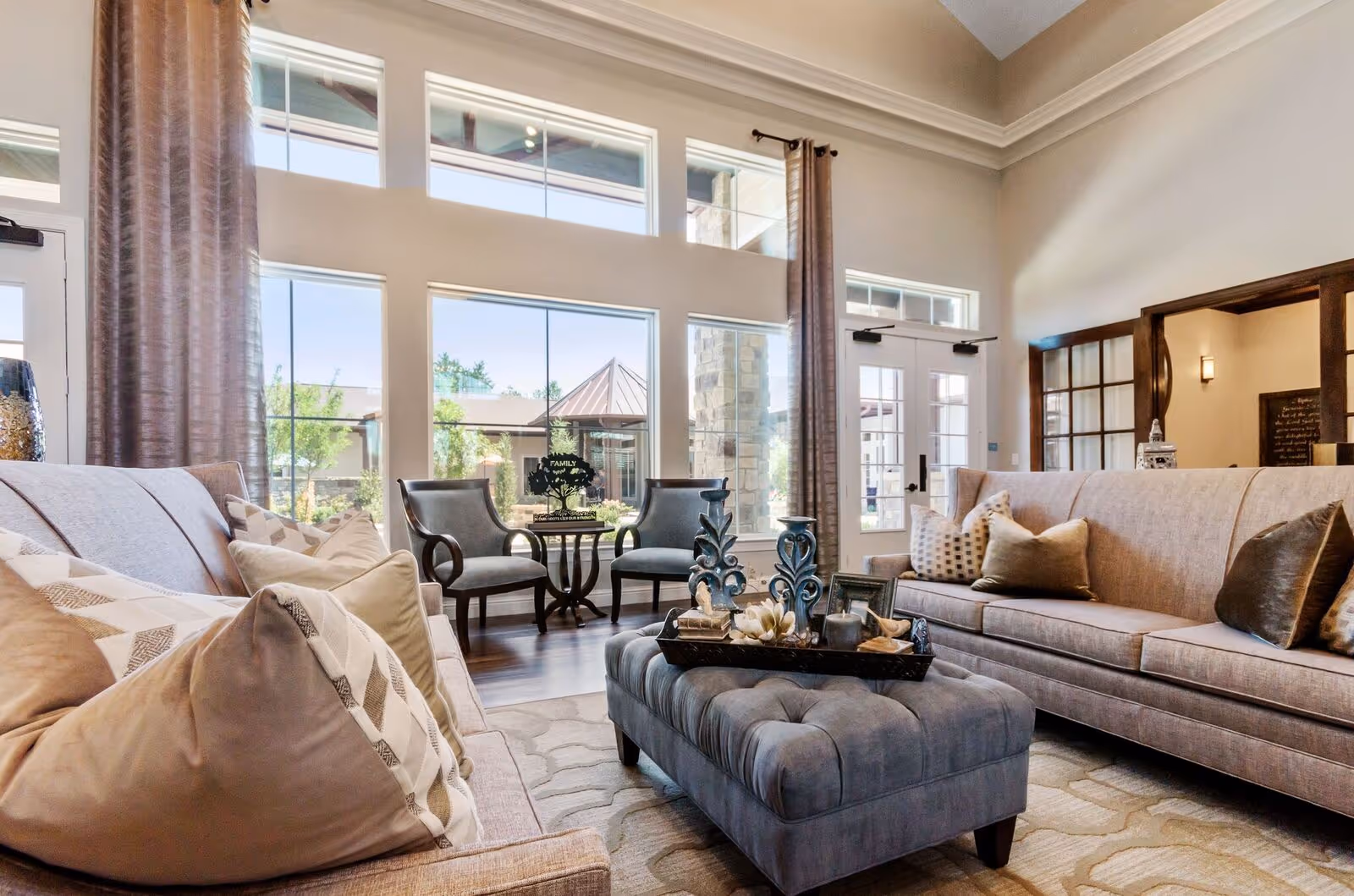 A bright and spacious living room with large windows letting in natural light. The room features two beige sofas with multiple decorative pillows, a tufted gray ottoman with a tray holding decorative items, two gray armchairs with wooden arms, and tall curtains framing the windows. Outside the windows, greenery and part of the building exterior are visible.
