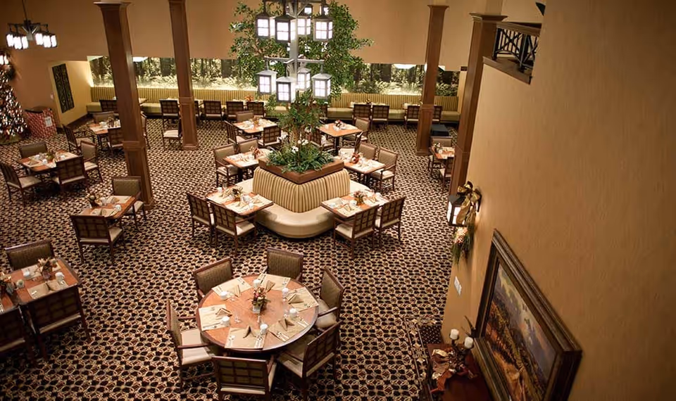Spacious senior living dining room with multiple round and square tables, a central planted booth, patterned carpet and hanging light fixtures.