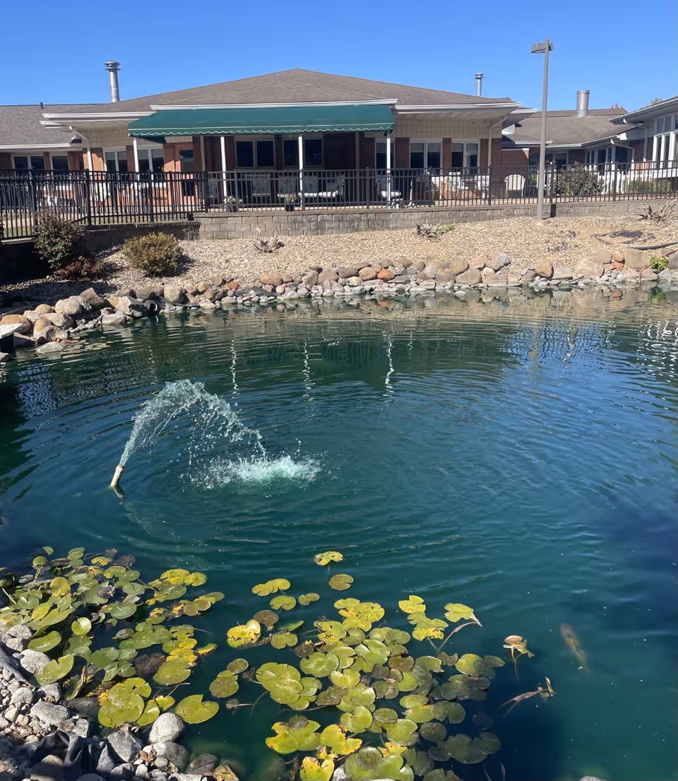 A pond with green lily pads and a small water fountain spraying water upwards in front of a senior living facility building with a green awning and a fenced patio area.