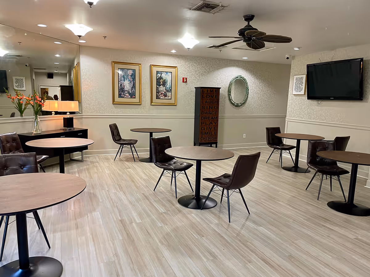 Small communal dining/activity room with round tables, brown chairs, a wall-mounted TV and decorative artwork.