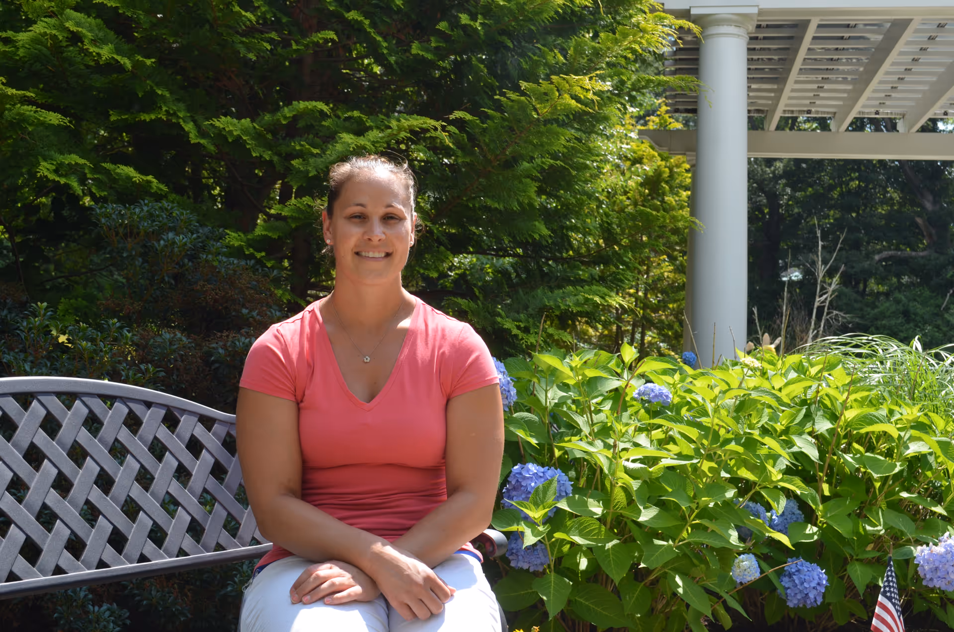 A woman wearing a pink shirt and white pants is sitting on a metal bench in a garden area with green bushes and purple flowers. There is a white pergola structure and trees in the background.