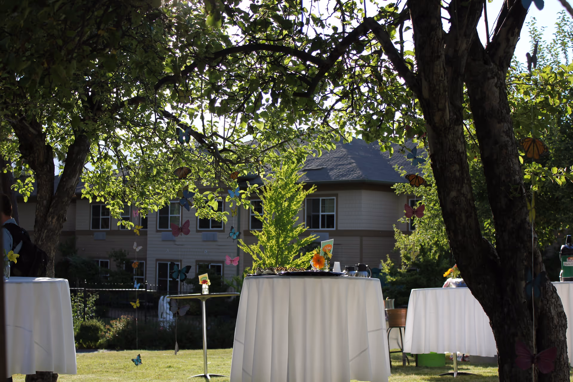 Outdoor garden area at Orchard Ridge Senior Living with round tables covered in white tablecloths set up on the grass. Trees with green leaves and hanging colorful butterfly decorations provide shade. A building with multiple windows is visible in the background.
