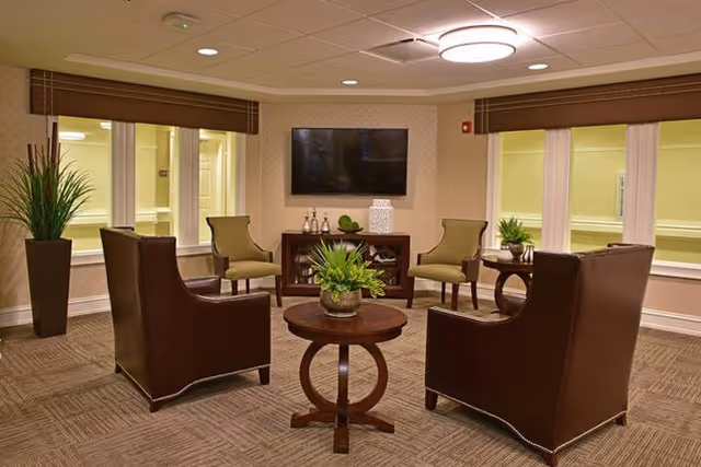 A cozy senior living facility lounge area with four brown leather armchairs and two green upholstered chairs arranged around two round wooden tables with potted plants. A flat-screen TV is mounted on the wall above a wooden cabinet with decorative items. The room has beige walls, carpeted floor, and windows with brown valances.