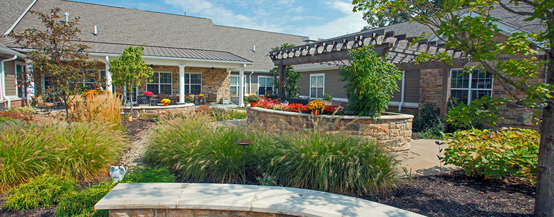 Outdoor garden area at Bickford of Carmel featuring a stone bench in the foreground, ornamental grasses, colorful flowers, a pergola with climbing plants, and a building with multiple windows and a covered porch in the background under a partly cloudy sky.