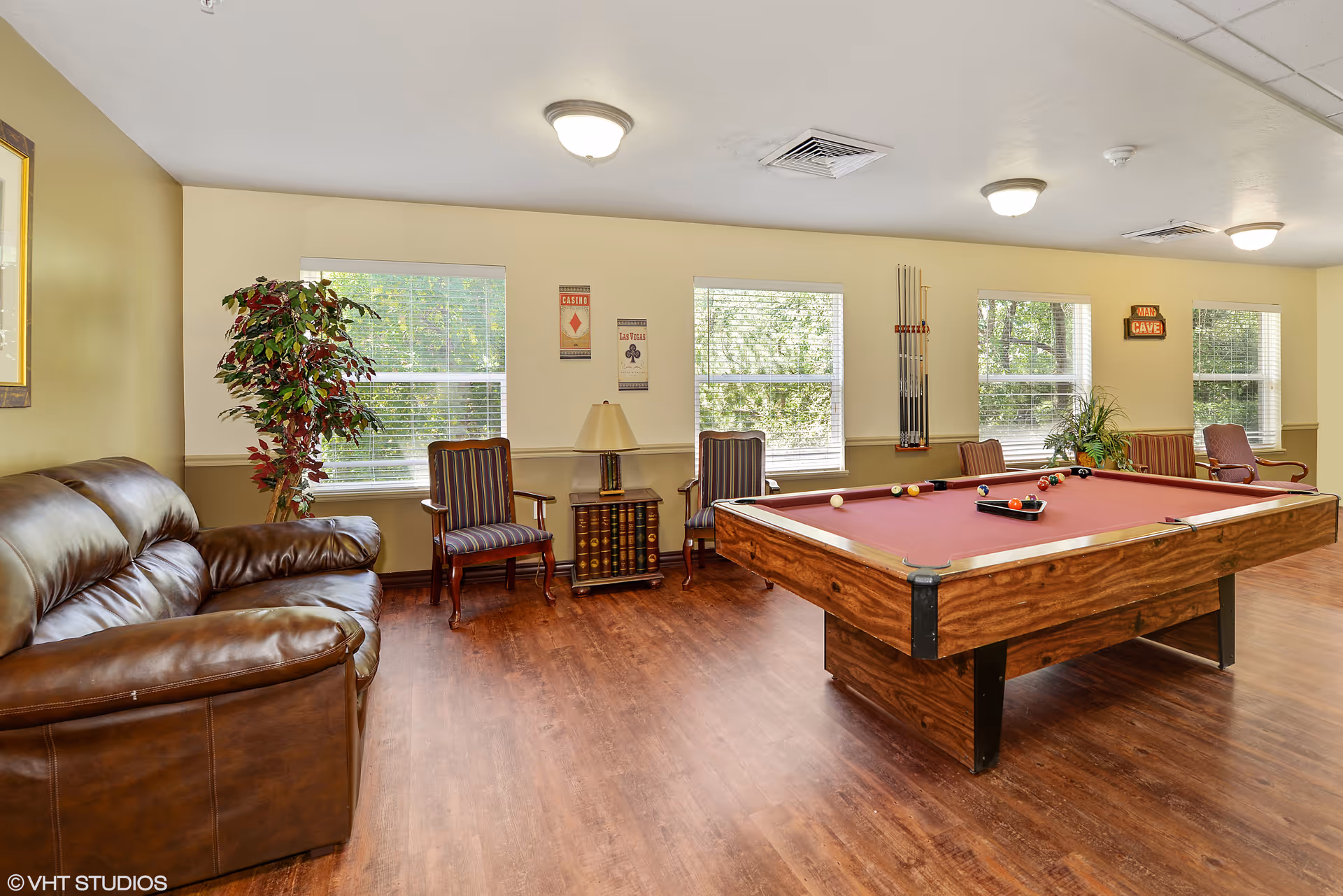 A recreational room with a brown leather couch on the left, two striped armchairs with a small table and lamp between them, and a pool table with a red felt surface on the right. The room has wooden flooring, several windows with white blinds, and wall decorations including a 'Man Cave' sign and casino-themed posters. Pool cues are mounted on the wall.