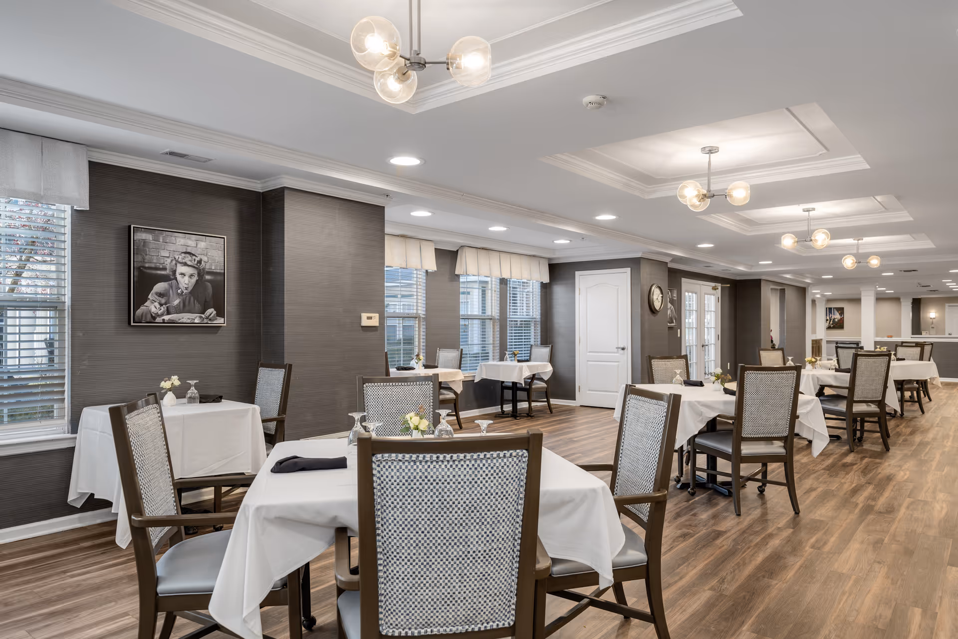A spacious dining room with multiple tables covered in white tablecloths, each set with glassware and small floral centerpieces. The room features wooden flooring, gray textured walls, large windows with white blinds and valances, and modern ceiling light fixtures. There is a black and white photo on one wall and a clock on another. The overall ambiance is clean, bright, and inviting.