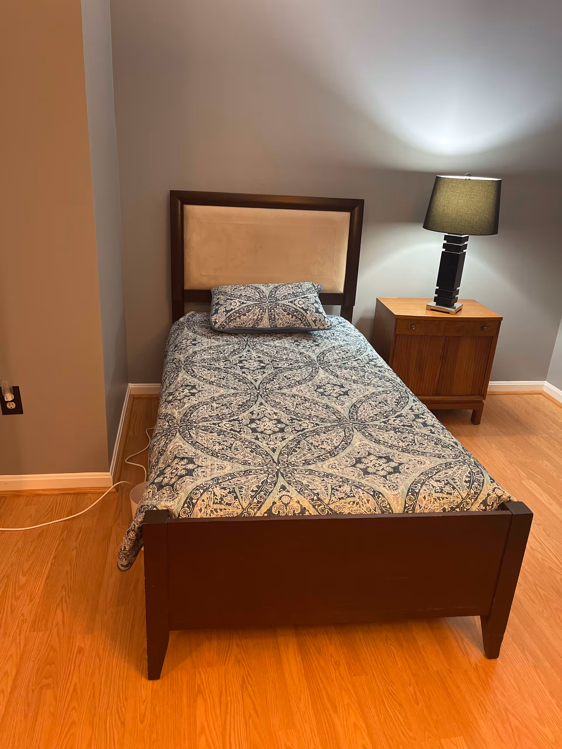 A single bed with a patterned blue and white bedspread and matching pillow, a wooden headboard with a beige cushioned center, a wooden nightstand with two drawers, and a black table lamp with a dark lampshade. The room has light wood flooring and gray walls.